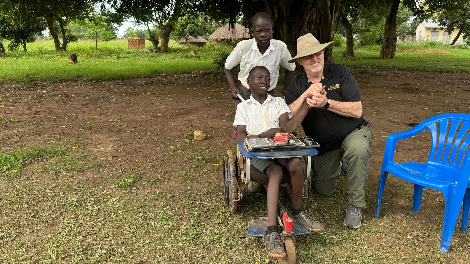Man in hat helps boy in wheelchair, another boy watches. Outdoors, grass, blue chair.