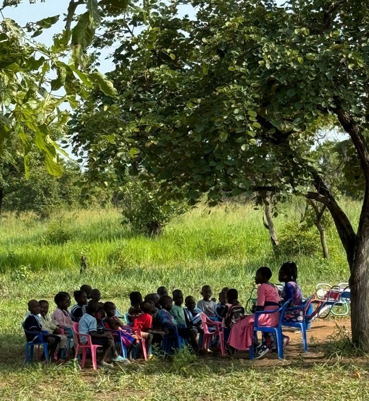 Children seated outdoors listening to two adults under a tree, surrounded by grass and foliage.
