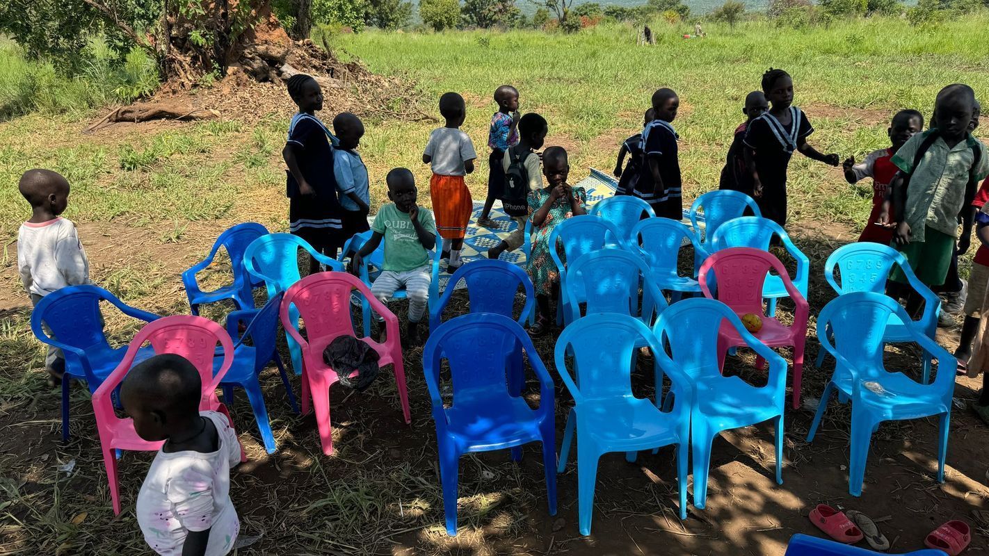 Children gathered around blue and pink plastic chairs outdoors, some are standing.