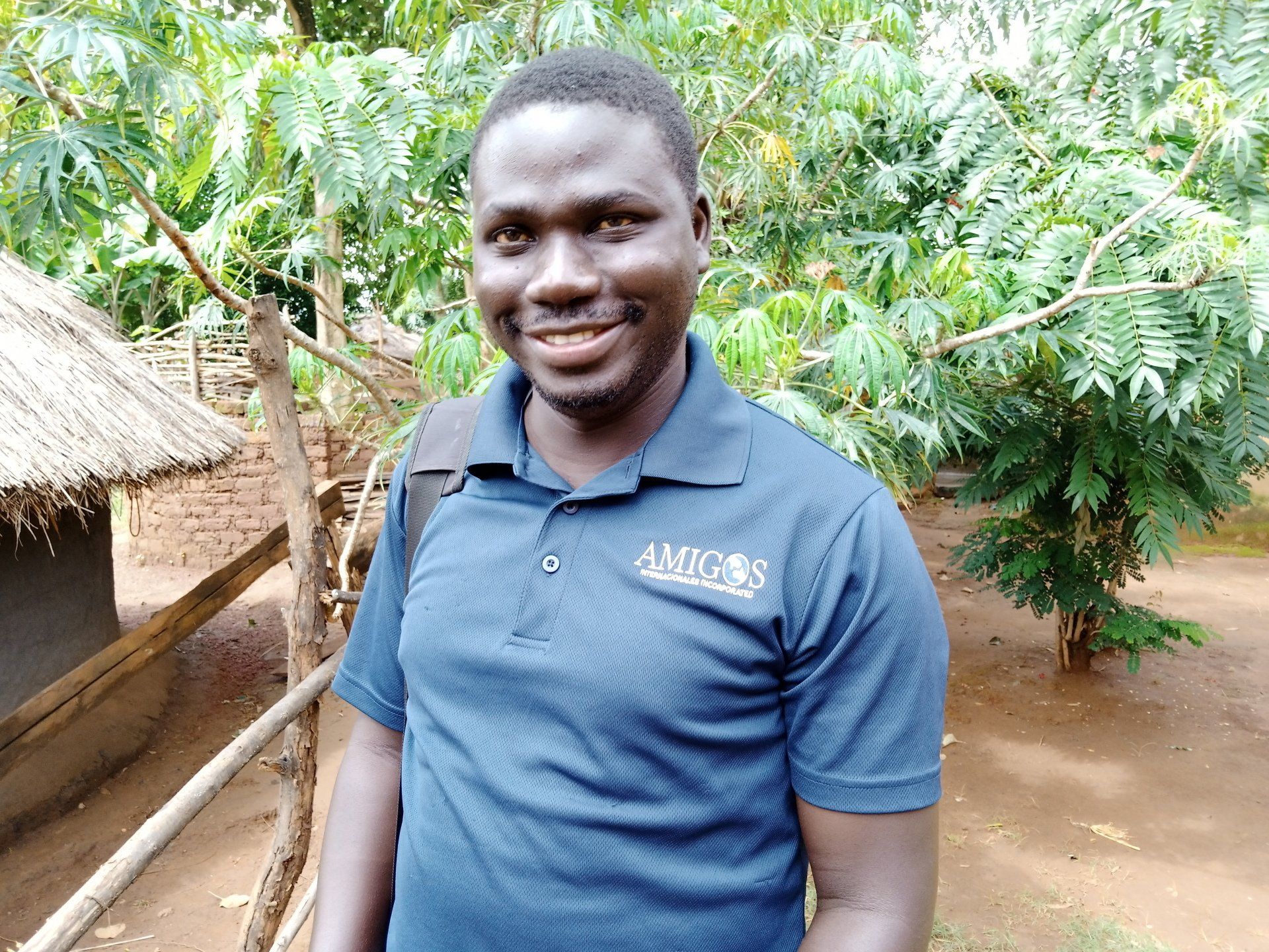 A man in a blue shirt is standing in front of a thatched hut.