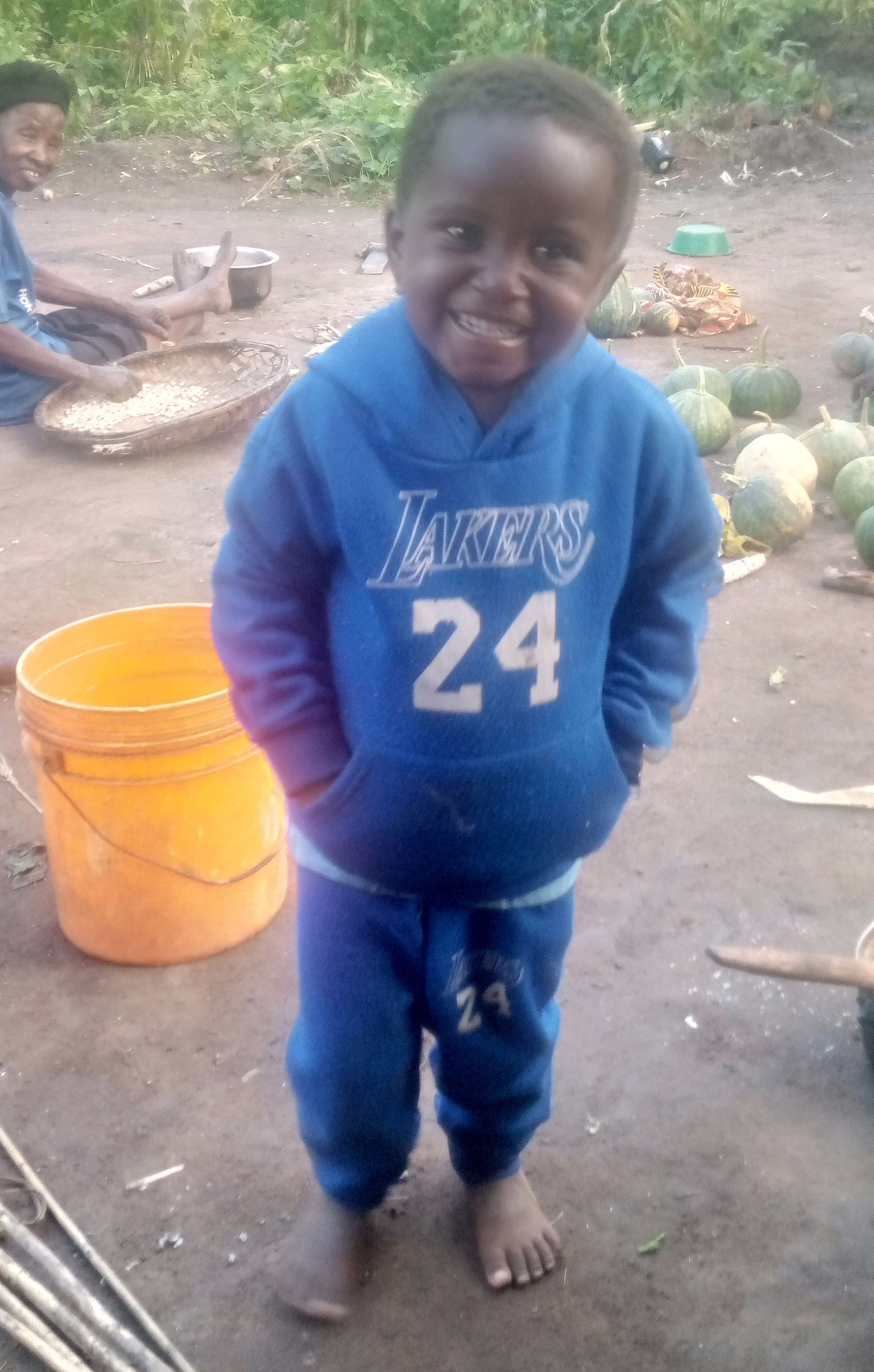 Young Black child wearing a blue Lakers hoodie and sweatpants, smiling, standing barefoot outdoors.