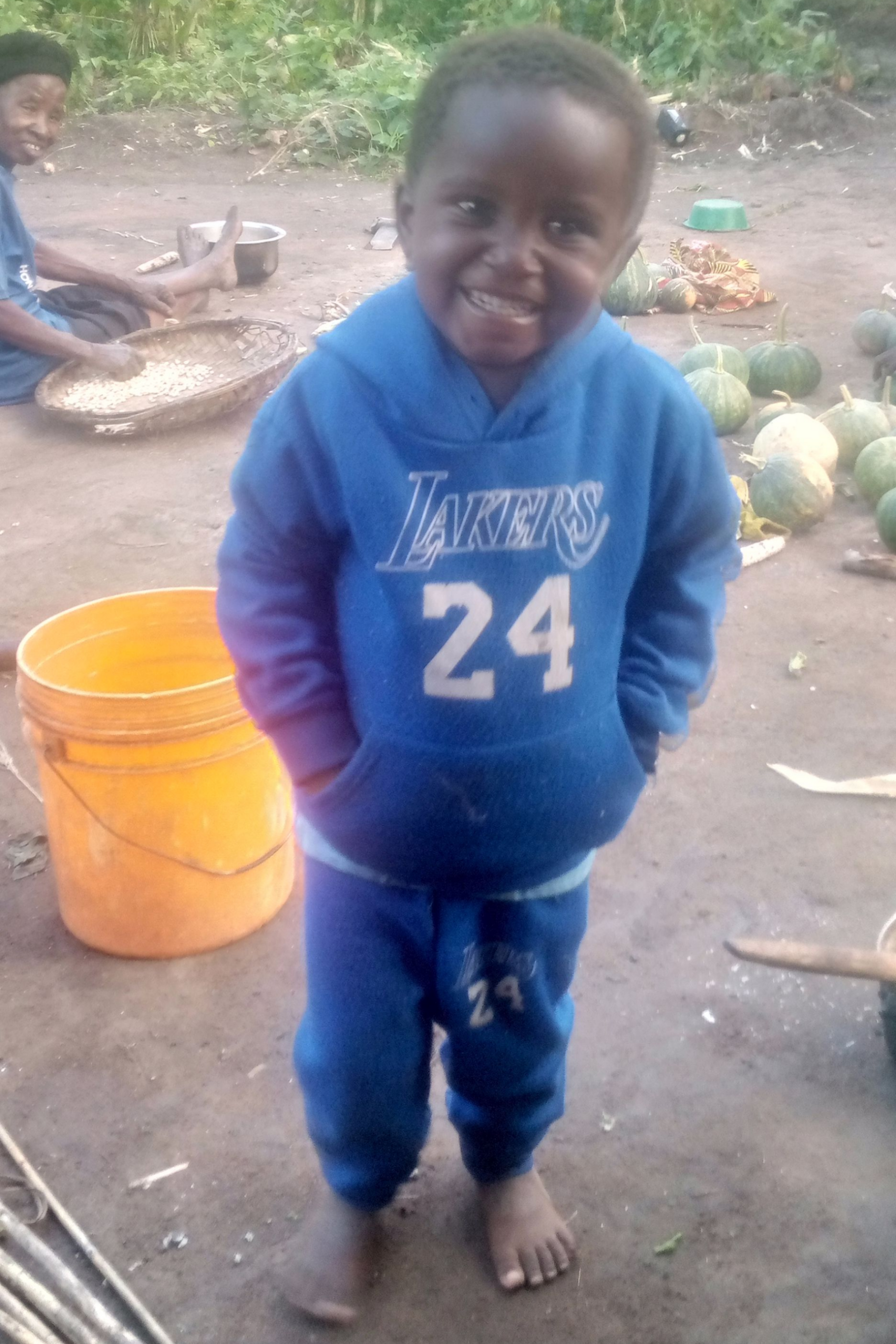 Young Black child wearing a blue Lakers hoodie and sweatpants, smiling, standing barefoot outdoors.