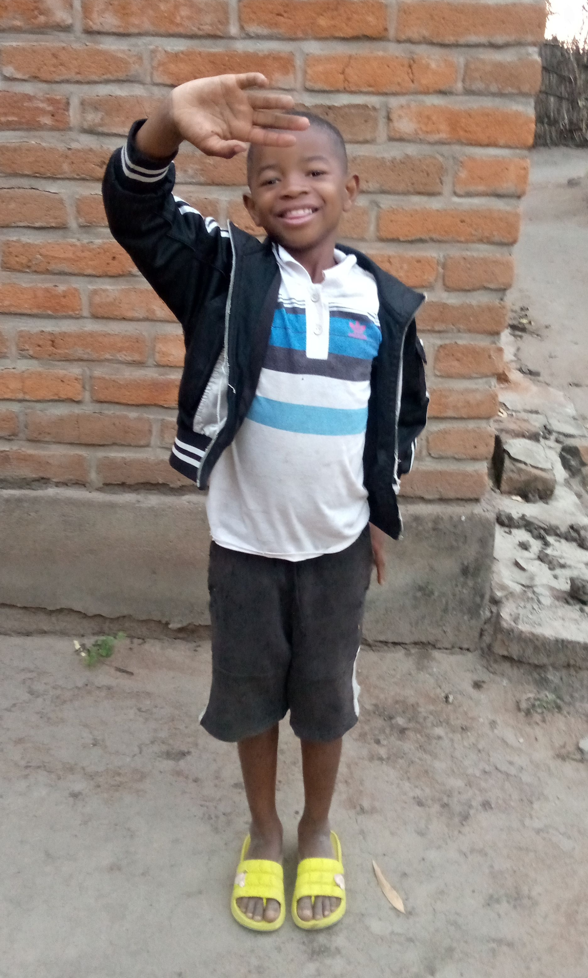 Smiling boy in black jacket and shorts waves, wearing yellow sandals, brick background.