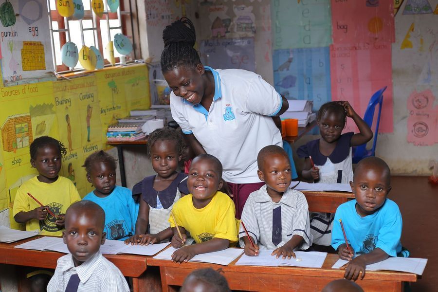 Teacher smiling at students in a colorful classroom; children at desks with pencils and paper.