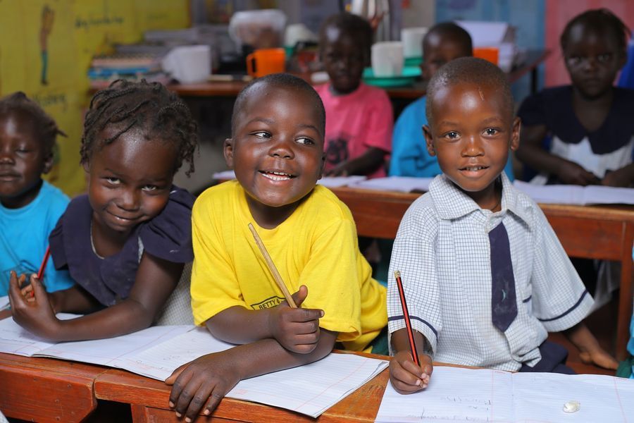 Children smiling, holding pencils, at desks in a brightly lit classroom.