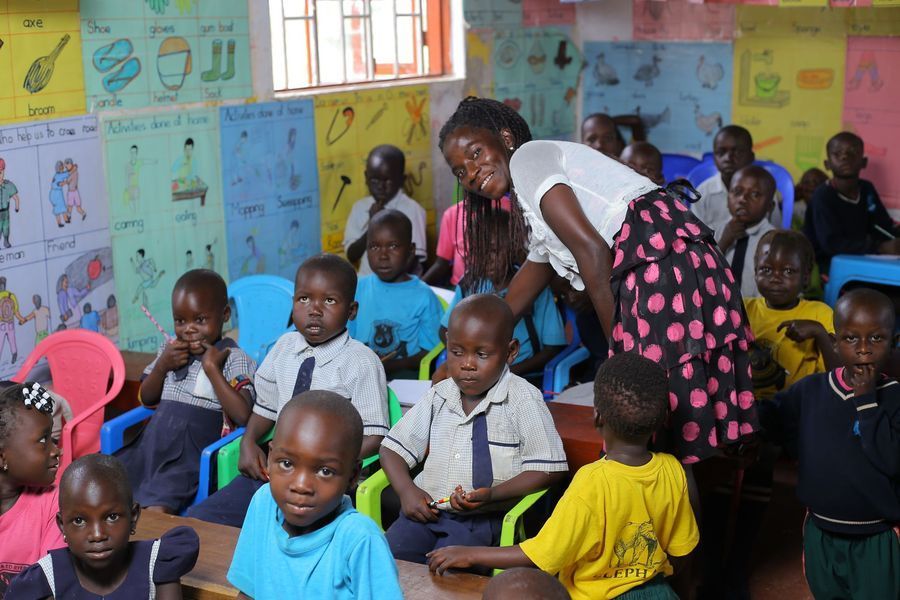 A teacher assists children in a colorful classroom, many in uniforms. Posters are on the wall.