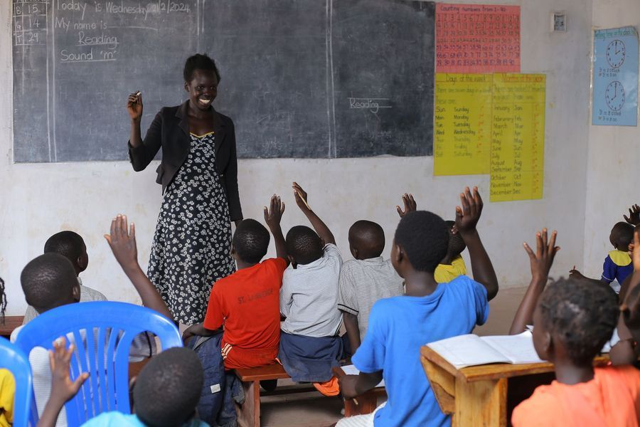 Teacher at blackboard with students raising hands in classroom.