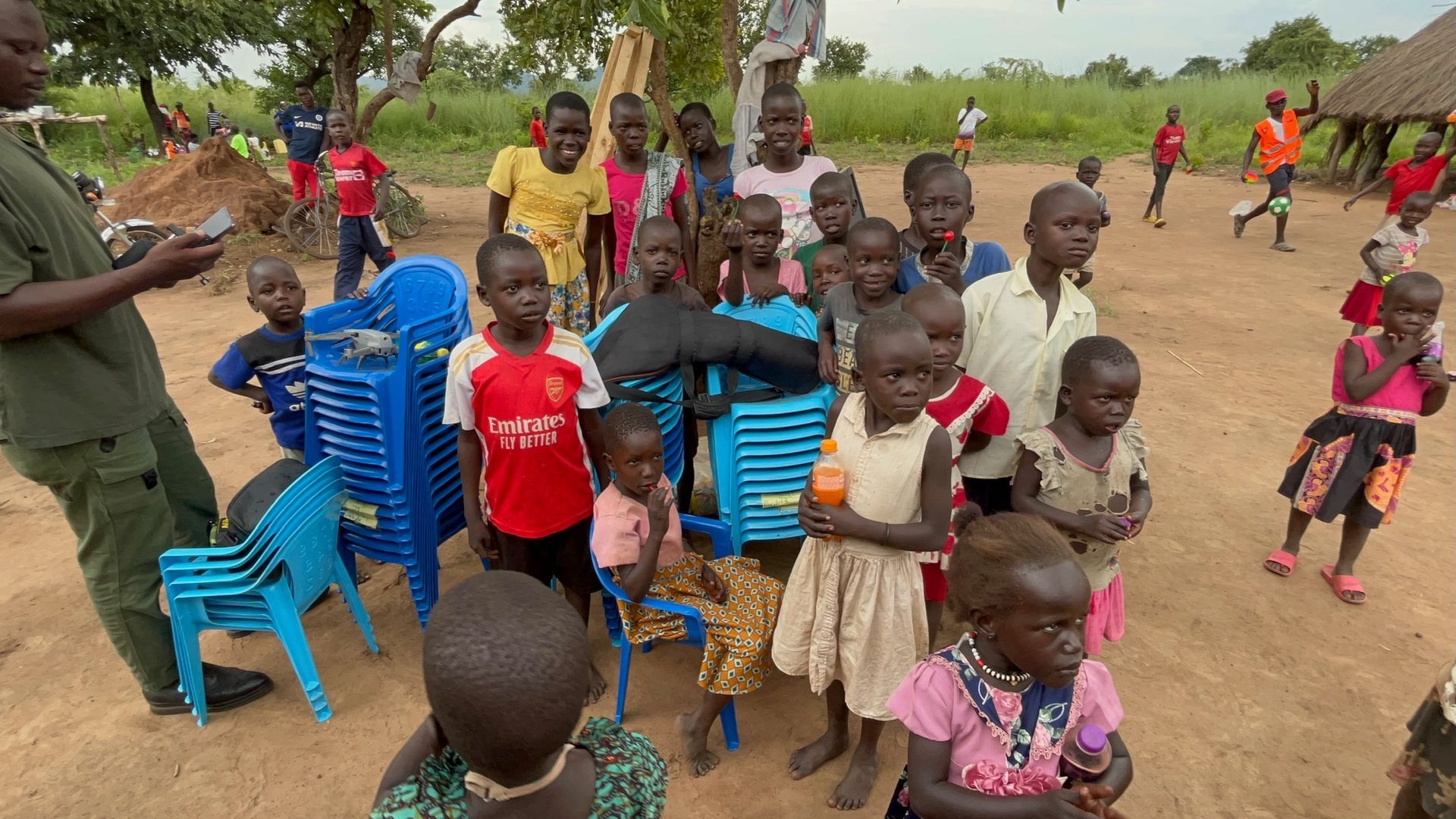 A group of children are standing around chairs in a dirt field.