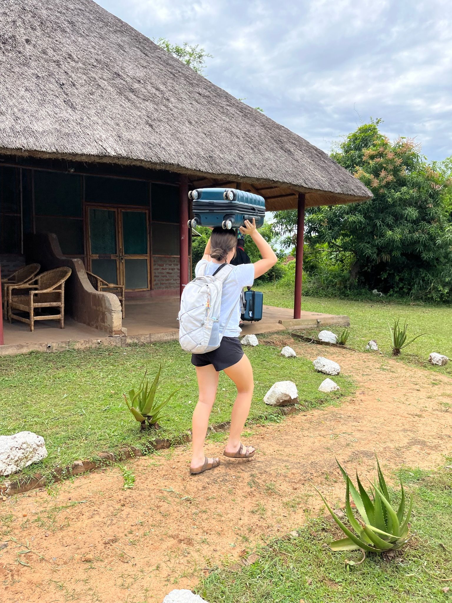 A woman is carrying a suitcase on her head in front of a thatched hut.