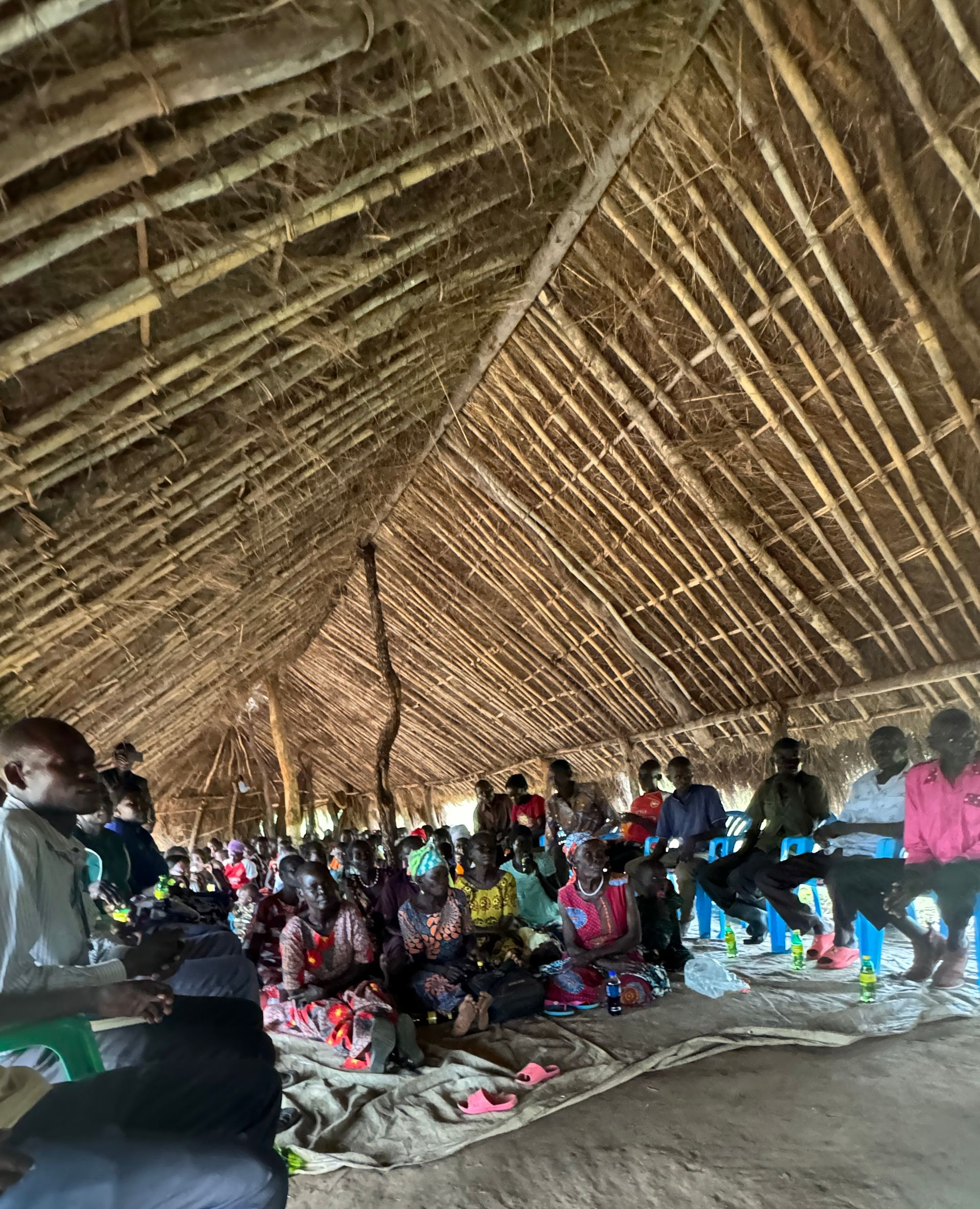People seated inside a thatched-roof shelter. They are listening to someone.