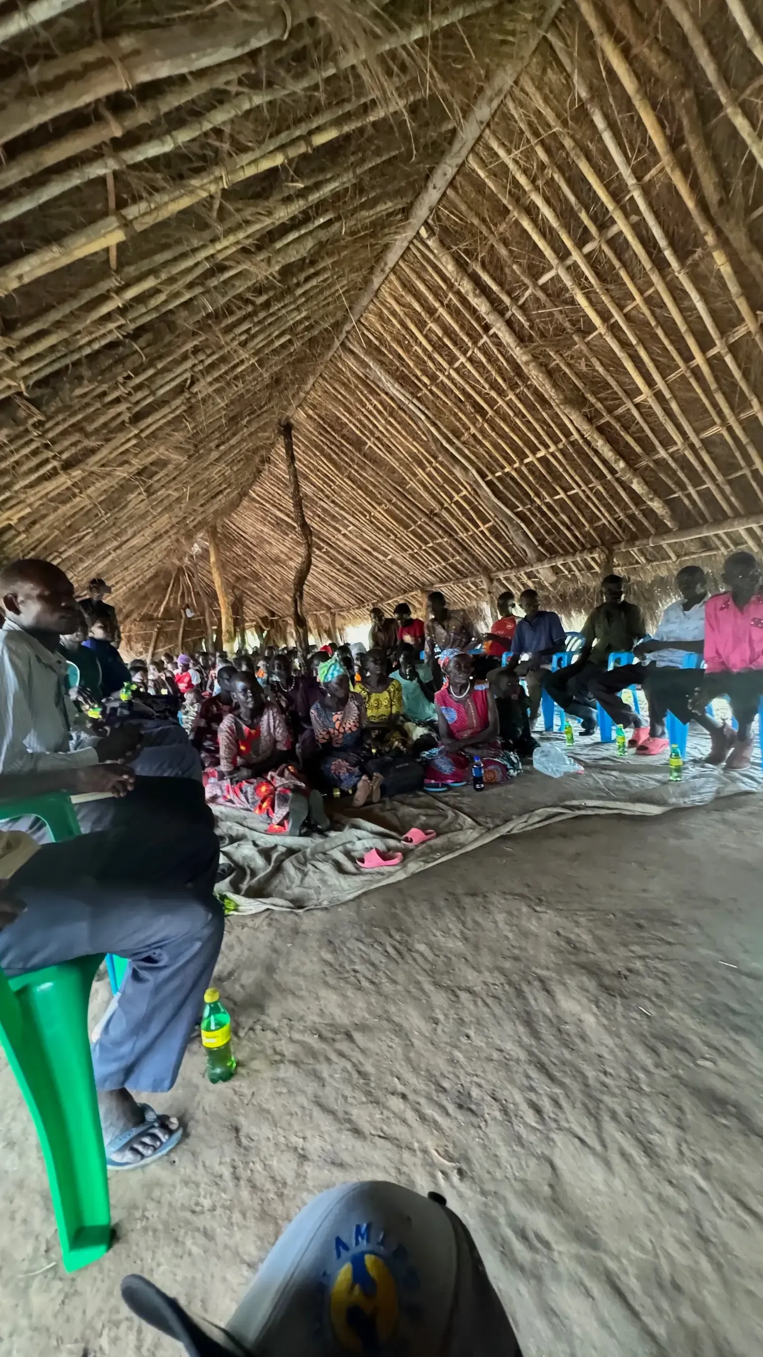 People seated under a thatched roof. Meeting or gathering. Daylight.