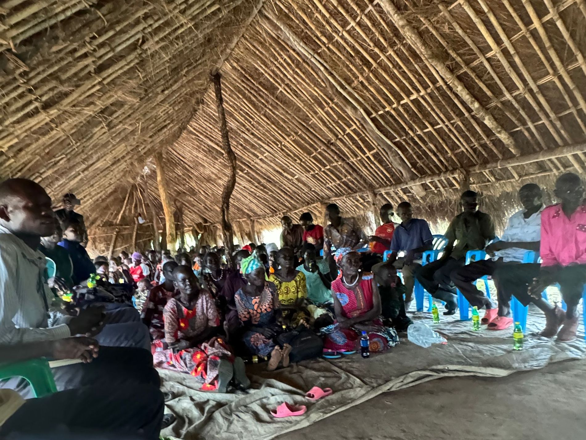 A large group of people are sitting in a hut under a thatched roof.