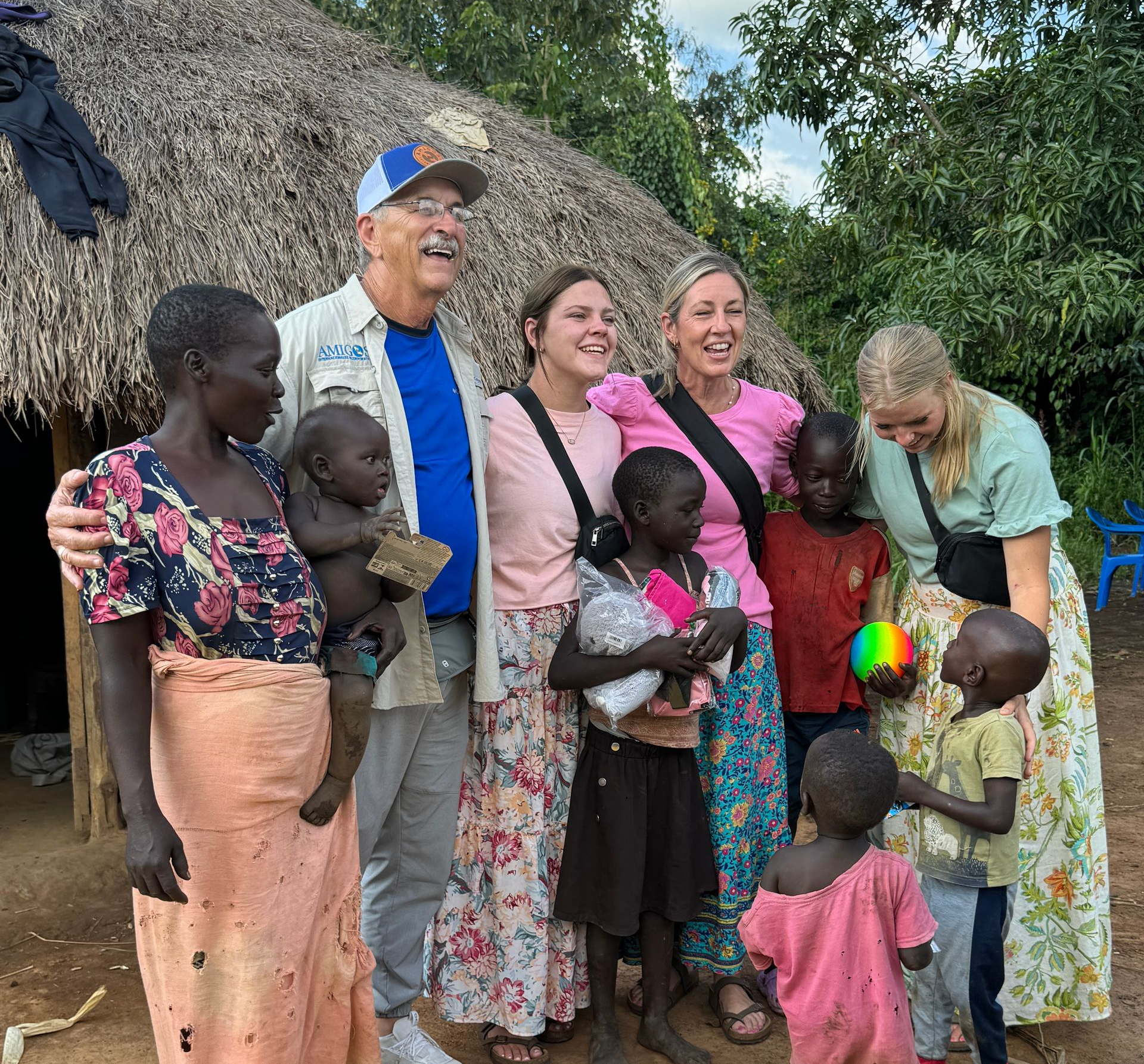 Group of adults and children smiling outside a thatched-roof structure.