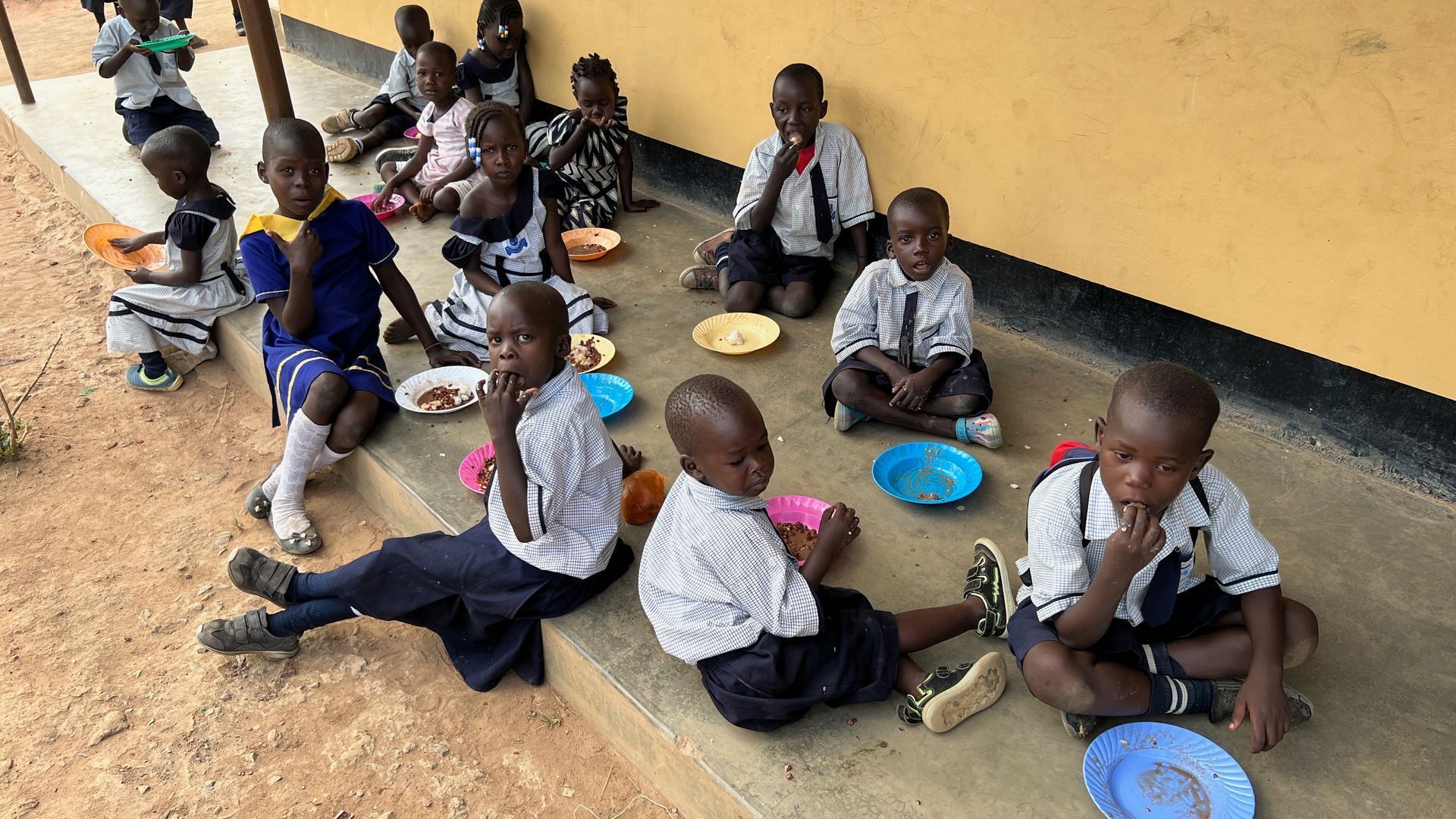 Children in uniforms sitting on a school porch, eating from small plates on the ground.