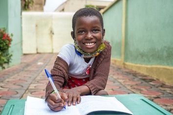 An Amigos sponsored young girl is sitting on the ground writing in a notebook . 
