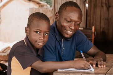 Amigos sponsors a man and a boy who are sitting at a table writing in a notebook .