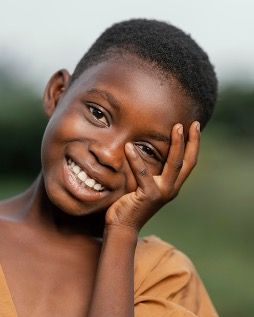 An Amigos sponsored young boy is smiling with his hand on his face .