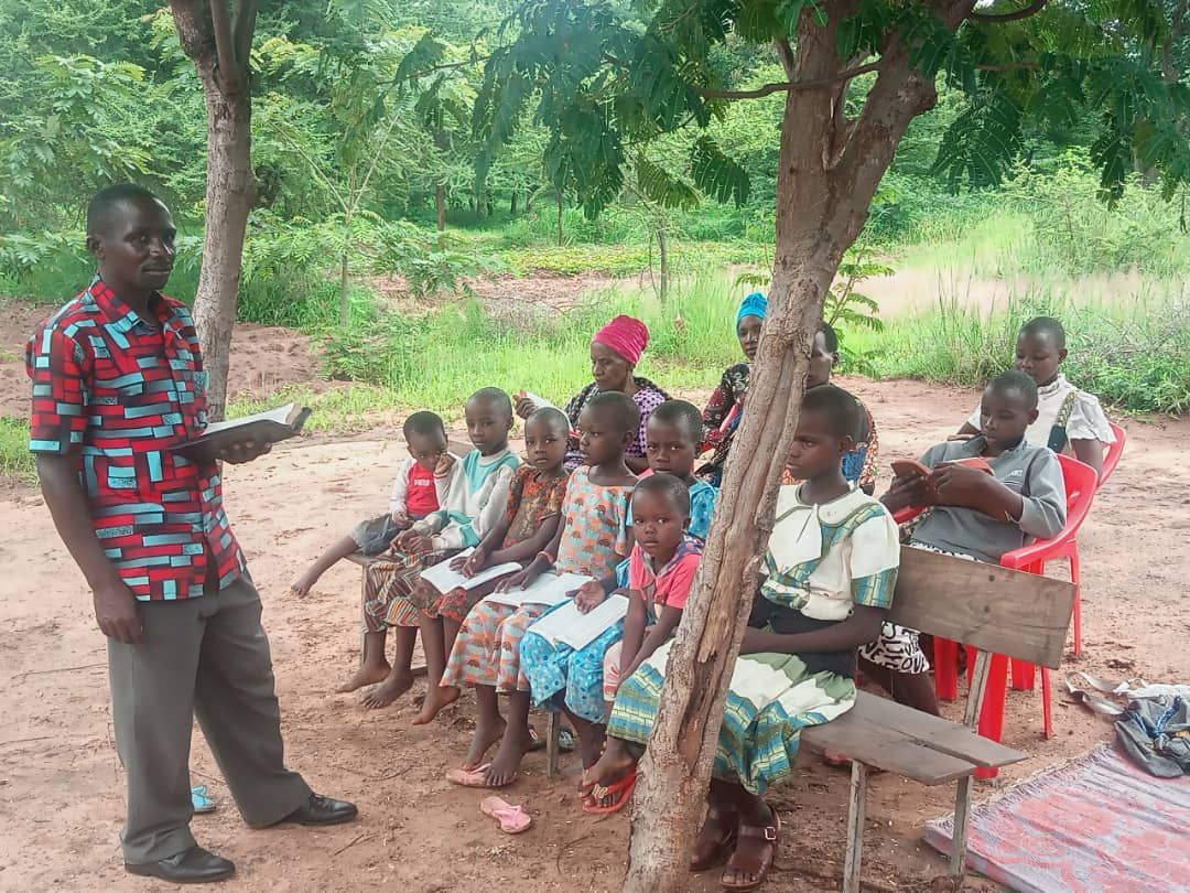 A man is standing in front of a group of children sitting under a tree.