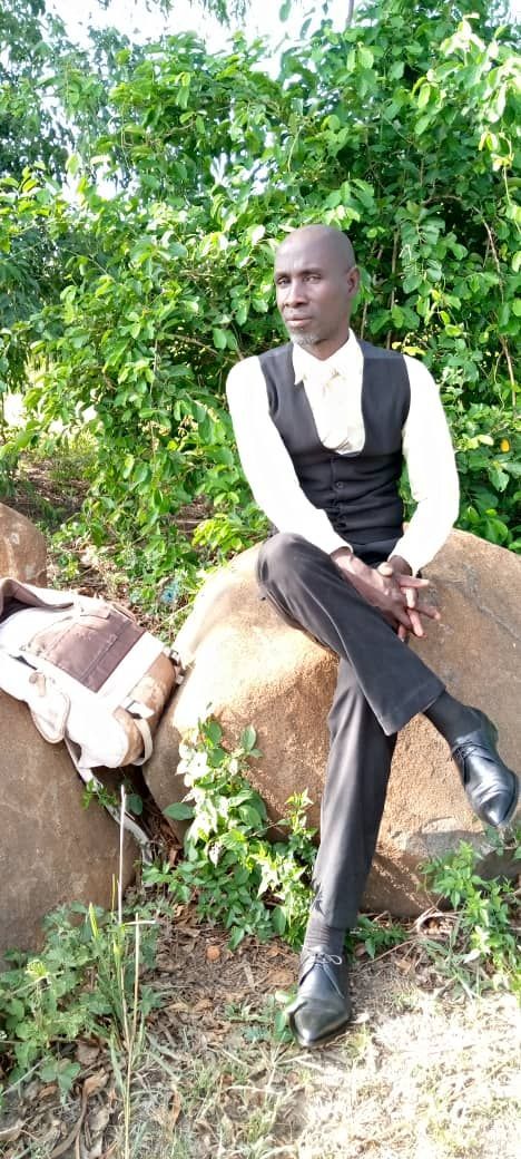 A person sits on a rock outside. They wear a vest, button-up shirt, and dress pants, looking away.