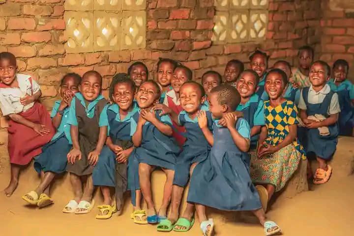 Group of children in blue school uniforms, laughing in a brick-walled room.