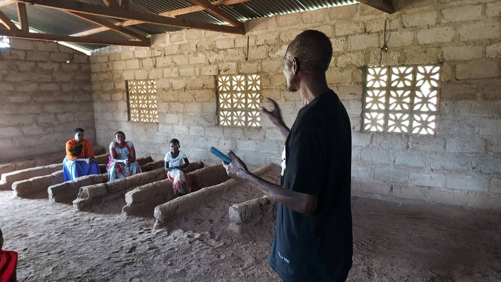 People, some in masks, at a health clinic. They are seated, with a healthcare worker checking a phone.