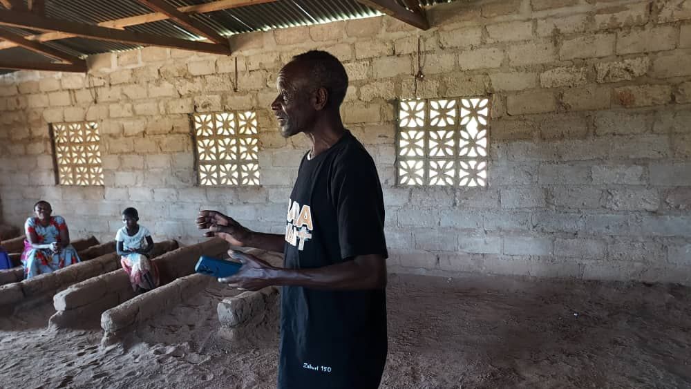 A person standing, facing a group of seated people in a room with dirt flooring.