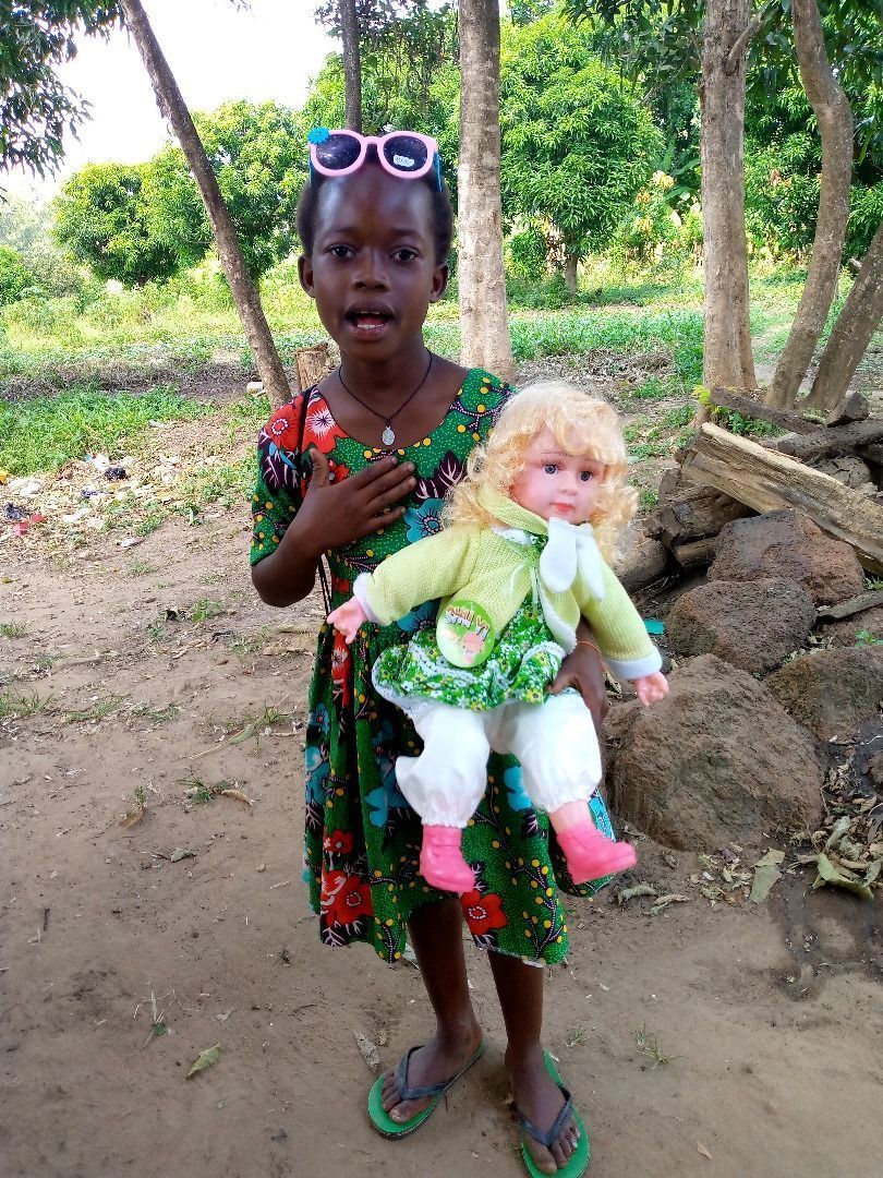 Girl holding doll, wearing sunglasses and green dress, standing outside.