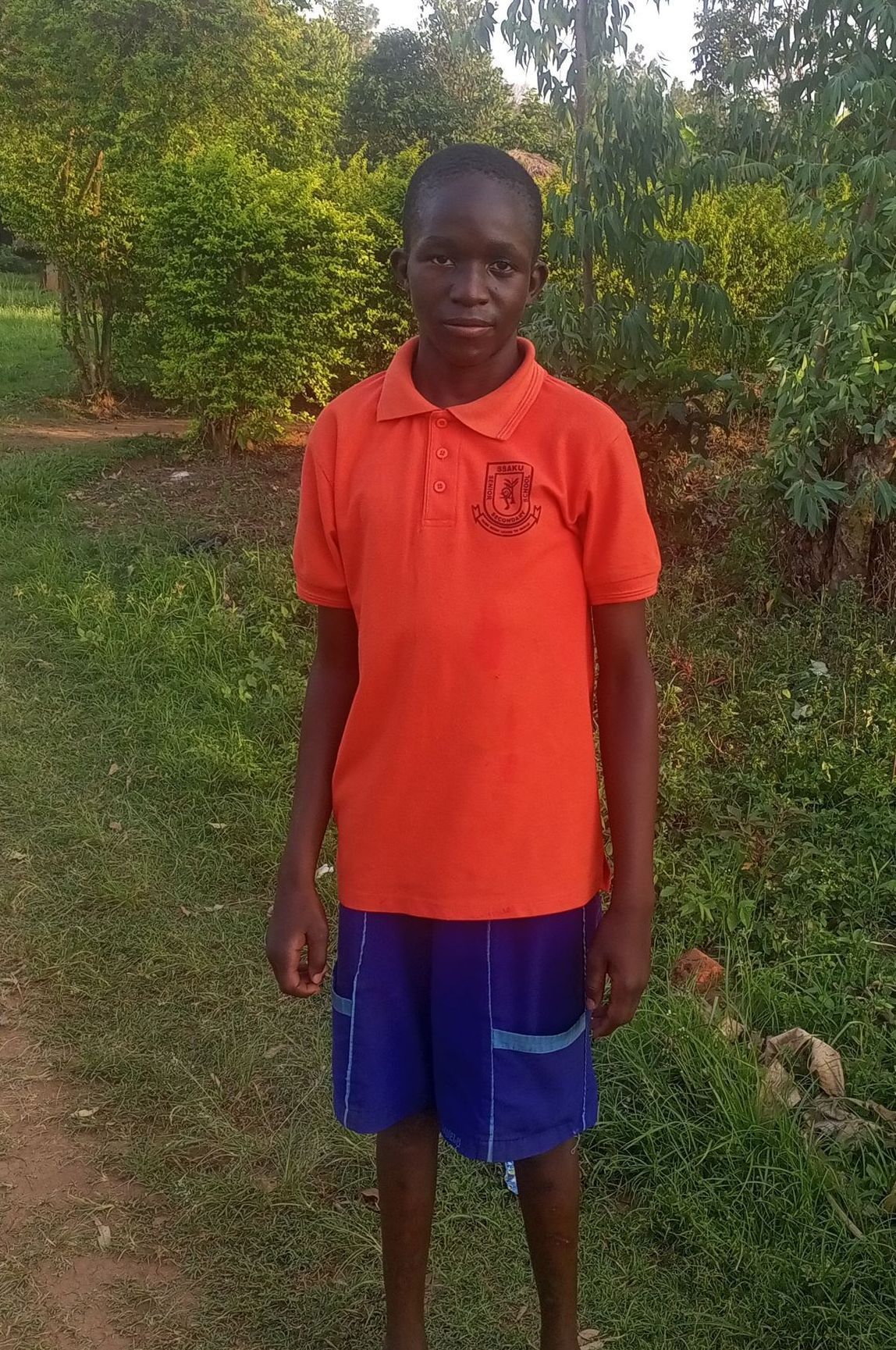A person in an orange shirt and blue shorts stands on a grassy path with a background of green foliage.