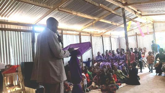 Man speaking to seated people in a simple building. Purple cloth, natural light.