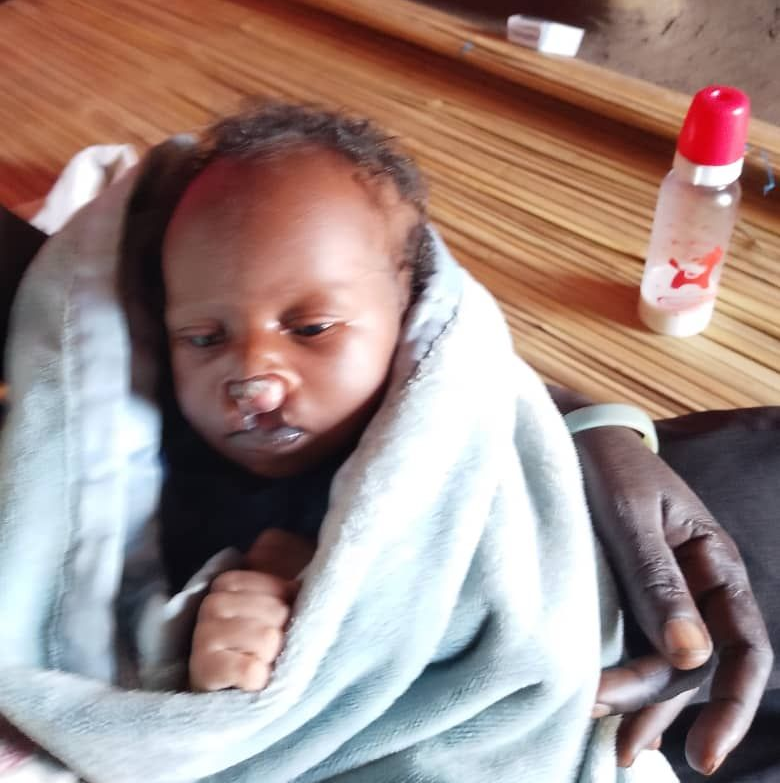 Baby swaddled in blue blanket, held by a person with dark skin, a bottle in the background.