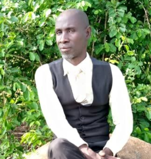 A person in a white long-sleeved shirt and black vest sits against a background of green foliage.