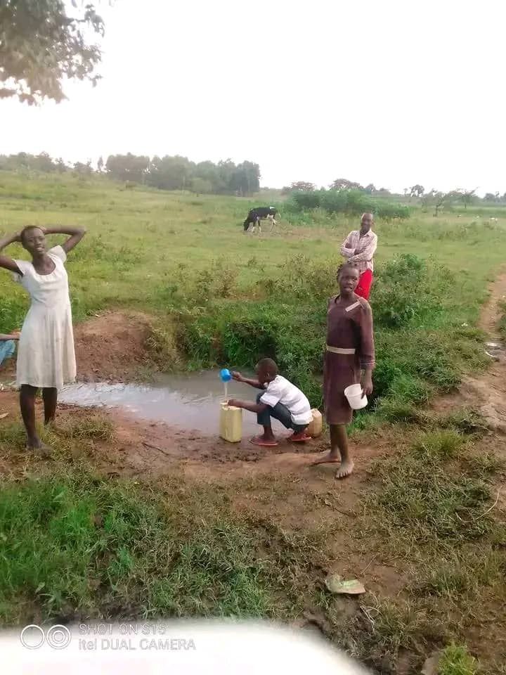 People gathering around a hand pump in a grassy rural field, with one person filling a container.
