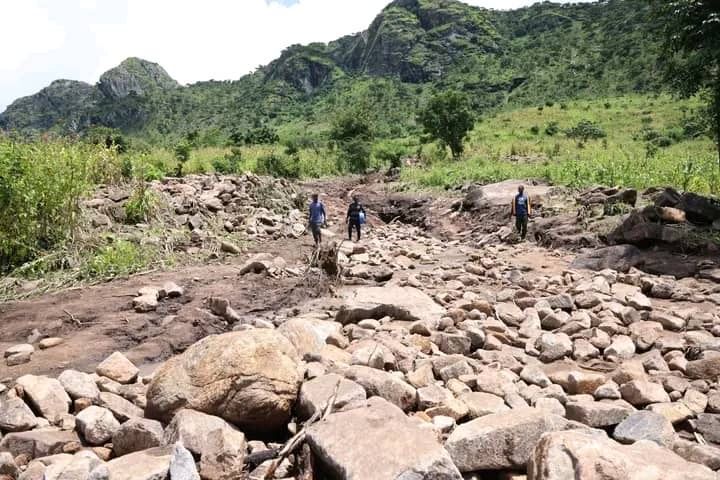 Three people walk through a dry, rocky riverbed surrounded by grassy hills and rocky mountains under a cloudy sky.