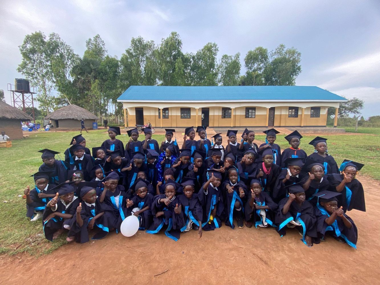 Children in graduation gowns pose for a photo in front of a school building.