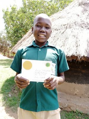 Young person smiles, holding certificate; green shirt, outdoors with thatched structure.