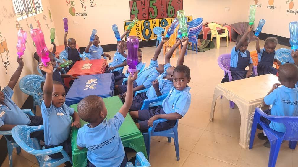 Children in blue shirts raise plastic bottles in a classroom with tables and colorful numbers on the wall.