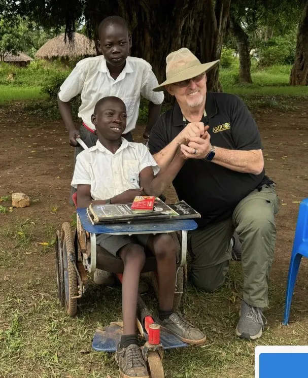 Two people beside a wheelchair outside, with a child seated at a small desk in a grassy outdoor area.