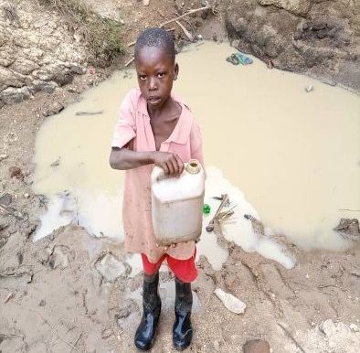 A young boy is standing in the mud holding a can of water.