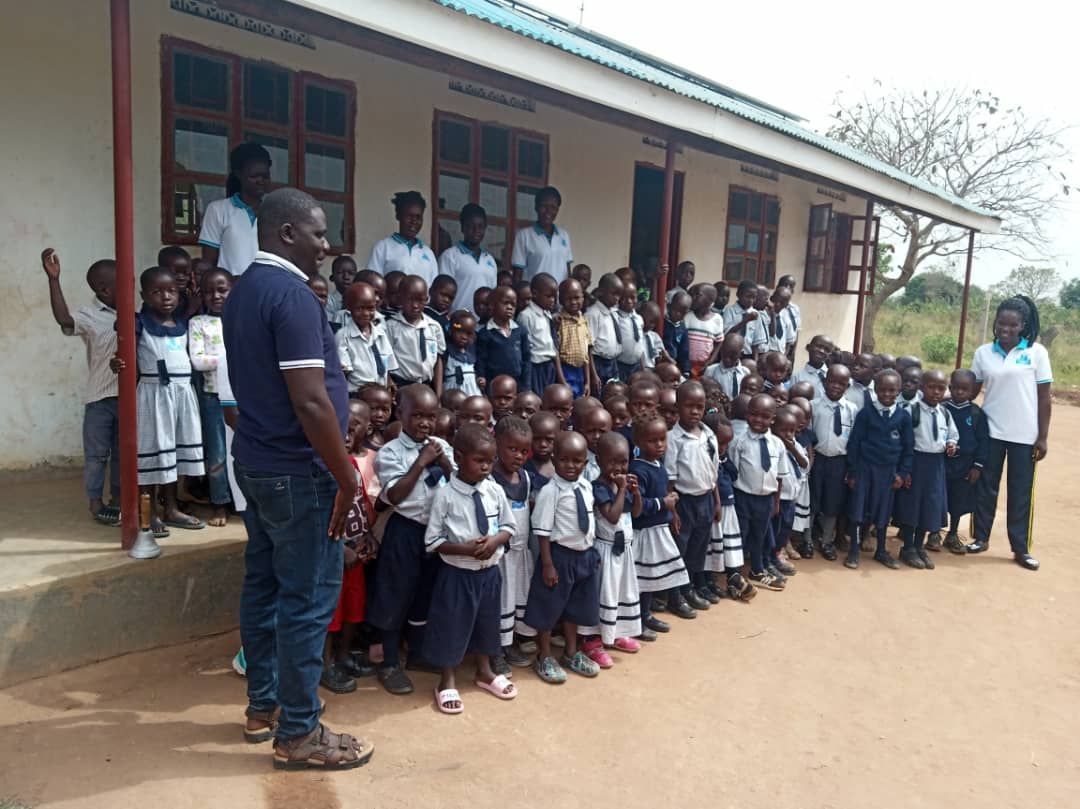 Children in school uniforms stand outside a building with teachers; sunlit, outdoors.
