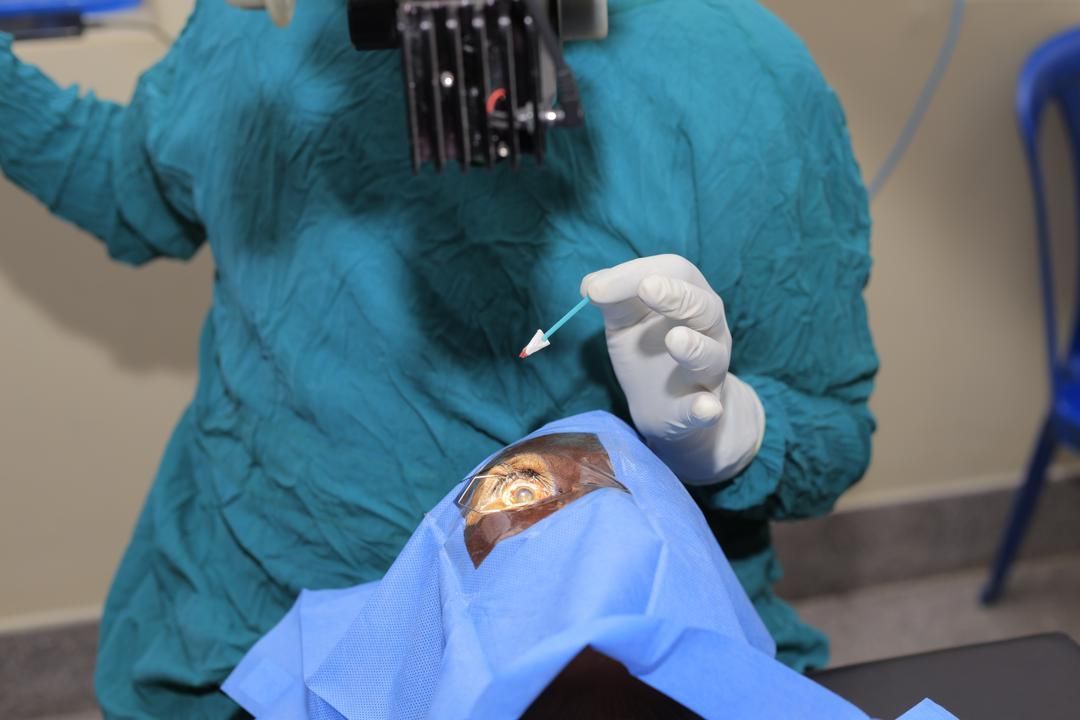 Surgeon in green scrubs operates on an eye. White-gloved hands hold a cotton swab.