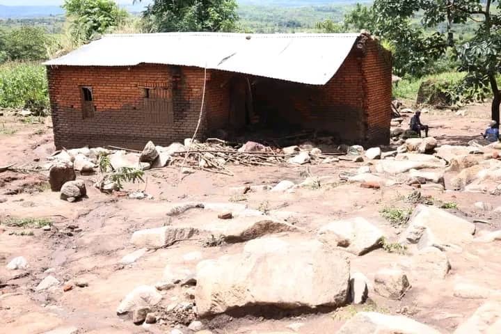 A small brick house with a metal roof stands surrounded by large rocks and debris in a rural landscape.