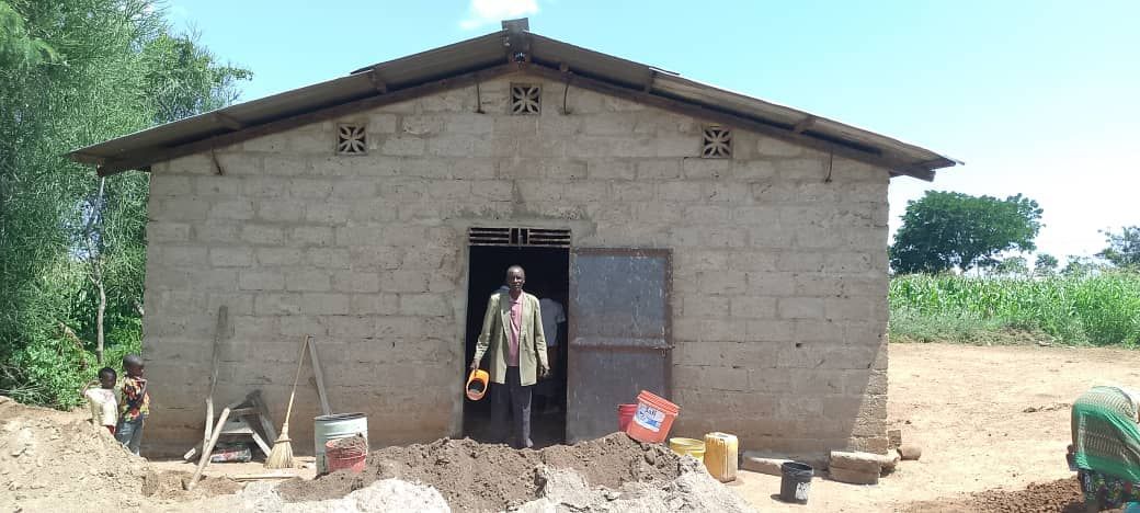 A man stands in the doorway of a mud brick building. Construction materials are in the foreground.