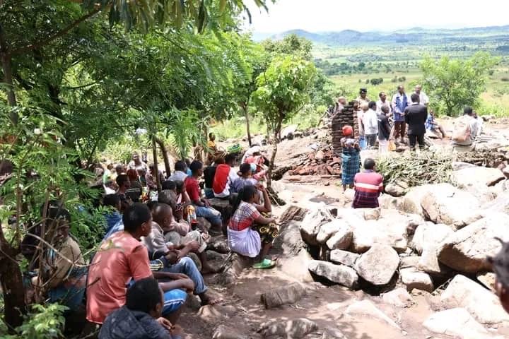 A group of people gathers outdoors in a rocky, rural landscape to attend a meeting or service held under a tree.