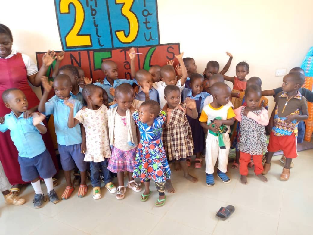 Children in classroom waving. Blue and white number sign in background.