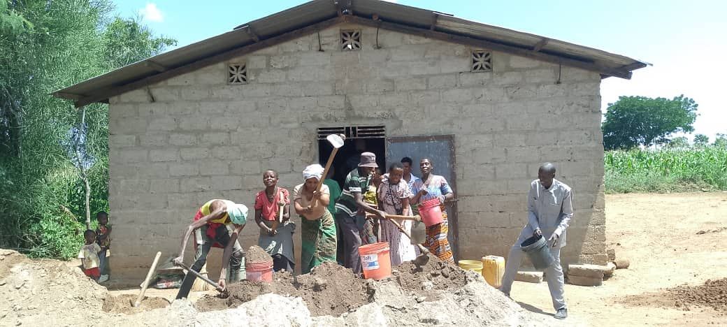 People working together in front of a building, some with shovels. Sunny outdoors.