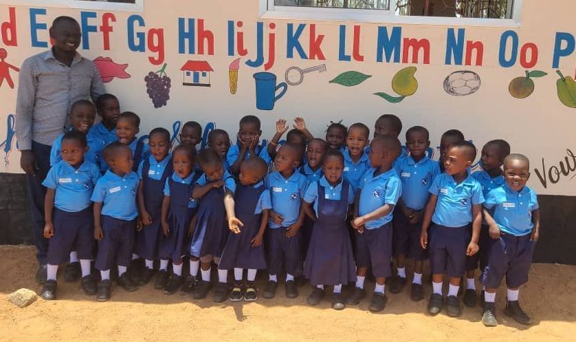 Teacher with a group of children in blue uniforms in front of a colorful alphabet mural.