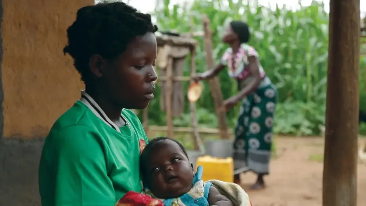 A person in a green shirt holds a baby outdoors, while another person works in a garden with tall corn stalks behind them.
