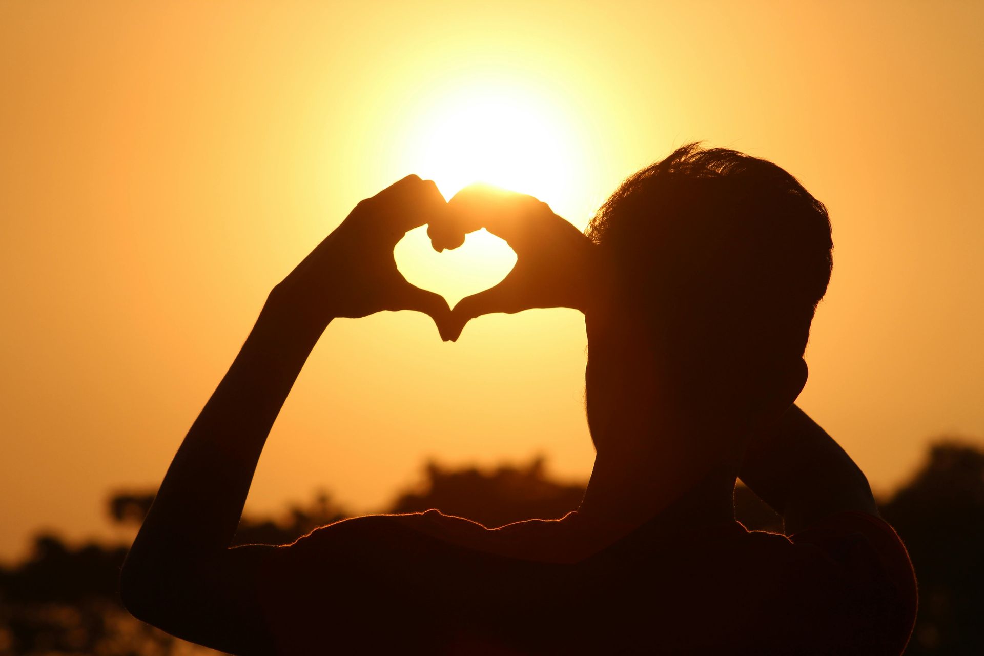 A person is making a heart shape with their hands in front of the sun