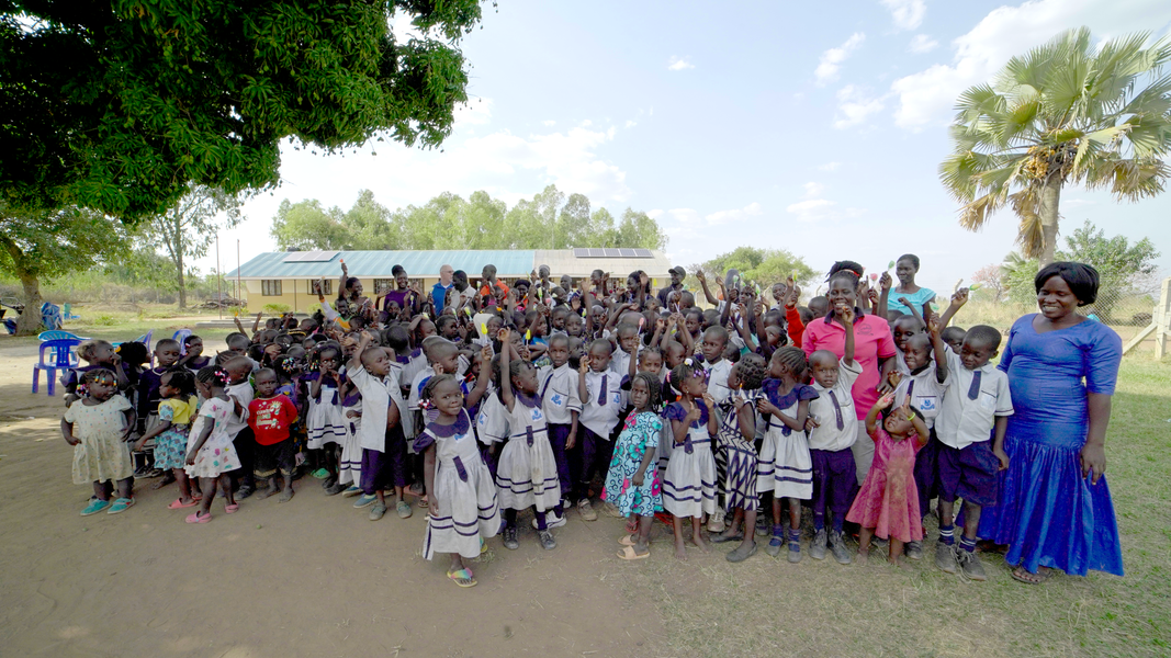 Group of children and adults standing outside a building, many raising their hands.