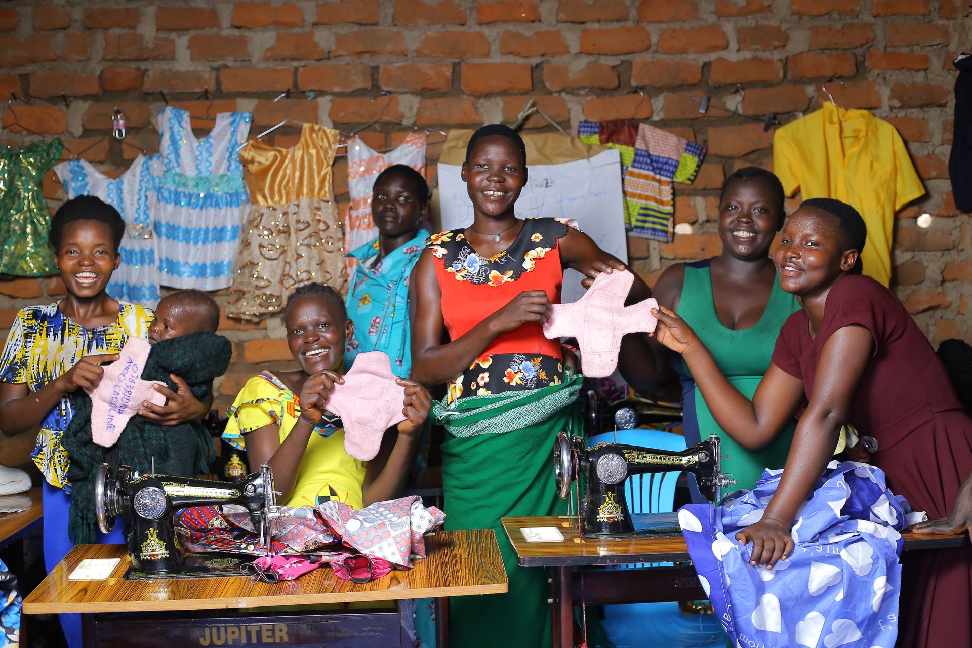 Group of women in sewing workshop, holding up handmade pads, smiling. Brick wall background, sewing machines present.